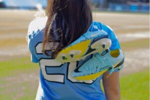Katie Thompson is looking at Dorrance Field in Chapel Hill wearing her blue UNC lacrosse jersey while holding her pair of blue and neon yellow Nike cleats over her shoulder.