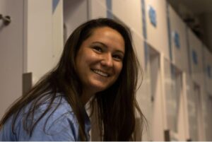 Katie Thompson is in the UNC women’s lacrosse locker room amongst a row of white lockers smiling and laughing during a conversation with her teammate Bailey Horne, who is unseen in the photo.
