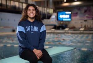 Aranza Vazquez sits on a teal diving board wearing a navy blue Carolina sweater. Behind her, swimmers are doing laps in a pool.