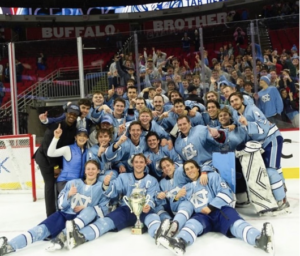 Photo of the UNC club hockey team, who are posing on the ice after winning the Governor’s Cup. The trophy is on the ice in the middle of the photo with the team, who is wearing light blue jerseys and navy shorts, surrounding it, with some players sitting on the ice, and some standing up. A group of fans is gathered behind the team, behind the glass posing for the picture with some celebrating. 